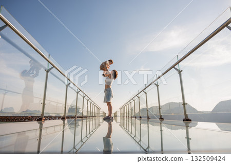 Caucasian woman holds her little son in her arms while standing on a glass bridge in the mountains. Caucasian woman holds her little son in her arms while standing on a glass bridge in the mountains. 132509424
