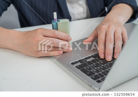 Business woman smoking an electronic cigarette while working on a laptop. Tobacco heating system. Business woman smoking an electronic cigarette while working on a laptop. Tobacco heating system. 132509654