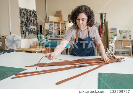 Caucasian woman makes belts from genuine leather in a workshop. Caucasian woman makes belts from genuine leather in a workshop. 132509767