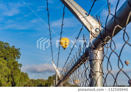Hot air balloon with a painted smile floats in a clear blue sky, viewed through barbed wire, creating a playful yet thought-provoking travel and freedom concept Hot air balloon with a painted smile floats in a clear blue sky, viewed through barbed wire, creating a playful yet thought-provoking travel and freedom concept 132509940