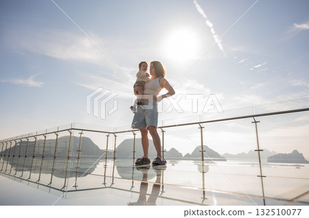 Caucasian woman holds her little son in her arms while standing on a glass bridge in the mountains. Caucasian woman holds her little son in her arms while standing on a glass bridge in the mountains. 132510077