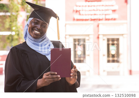 African woman in hijab and graduation gown holding diploma.  132510098
