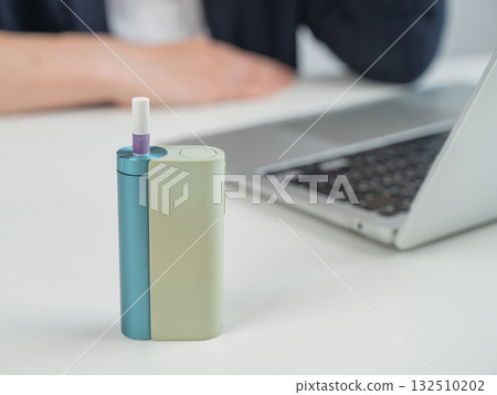 Business woman smoking an electronic cigarette while working on a laptop. Tobacco heating system in the foreground. Business woman smoking an electronic cigarette while working on a laptop. Tobacco heating system in the foreground. 132510202