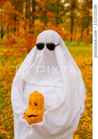 Woman in white sheet holding jack o'lantern against autumn forest background. Halloween costume. Vertical photo. Woman in white sheet holding jack o'lantern against autumn forest background. Halloween costume. Vertical photo. 132510286