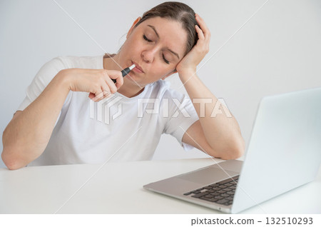 Tired woman smoking an electronic cigarette while working on a laptop. Tobacco heating system. Tired woman smoking an electronic cigarette while working on a laptop. Tobacco heating system. 132510293