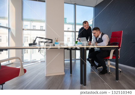 Caucasian man standing at colleague's desk discussing work. Caucasian man standing at colleague's desk discussing work. 132510340