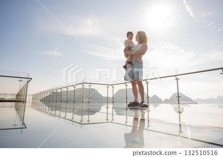 Caucasian woman holds her little son in her arms while standing on a glass bridge in the mountains. Caucasian woman holds her little son in her arms while standing on a glass bridge in the mountains. 132510516