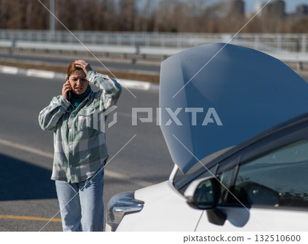 Caucasian woman cries and calls on the phone standing near the car with the hood open.  132510600