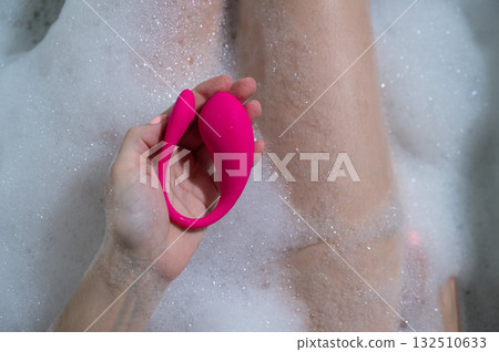 A woman lies in a bubble bath and holds a curved pink vibrator. A woman lies in a bubble bath and holds a curved pink vibrator. 132510633