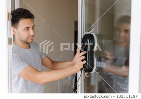 Happy young man installing robotic vacuum cleaner on window, preparing to activate device for efficient cleaning session, ensuring spotless interior environment. Cleaning house with smart devices. Happy young man installing robotic vacuum cleaner on window, preparing to activate device for efficient cleaning session, ensuring spotless interior environment. Cleaning house with smart devices. 132511187