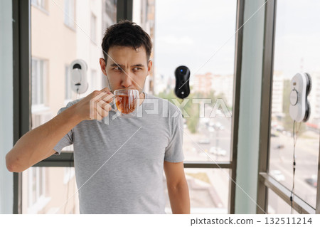 Relaxed young man in casual attire enjoying tea while automated window cleaning robots work in background. Modern smart home technology meeting everyday relaxation moments in urban setting. Relaxed young man in casual attire enjoying tea while automated window cleaning robots work in background. Modern smart home technology meeting everyday relaxation moments in urban setting. 132511214