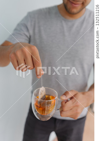 Close-up vertical cropped shot of young man dipping tea bag into glass cup filled with steaming hot water, crafting soothing and aromatic beverage perfect for relaxation and enjoyment indoors. Close-up vertical cropped shot of young man dipping tea bag into glass cup filled with steaming hot water, crafting soothing and aromatic beverage perfect for relaxation and enjoyment indoors. 132511216