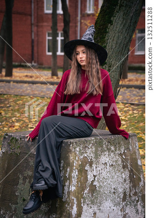 Teen girl in a witch hat and burgundy sweater sits on a stone surface surrounded by autumn leaves and trees near brick buildings 132511288
