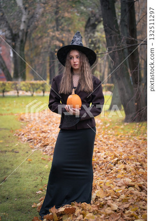 Teen girl in a witch hat holds an orange pumpkin close to the camera, creating a mystical autumn portrait with soft natural light Teen girl in a witch hat holds an orange pumpkin close to the camera, creating a mystical autumn portrait with soft natural light 132511297