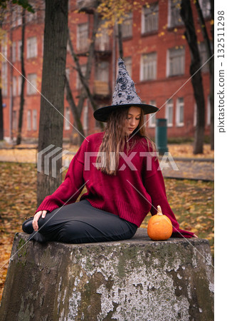Teen girl in burgundy sweater and witch hat holds a pumpkin outdoors, surrounded by autumn leaves and brick buildings, creating a cozy Halloween vibe 132511298