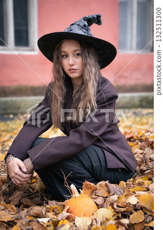 Teen girl in a witch hat and dark outfit sits among fallen leaves with a small pumpkin at her feet, framed by an old pink building wall 132511300