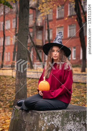 Teen girl in burgundy sweater and witch hat holds a pumpkin outdoors, surrounded by autumn leaves and brick buildings, creating a cozy Halloween vibe Teen girl in burgundy sweater and witch hat holds a pumpkin outdoors, surrounded by autumn leaves and brick buildings, creating a cozy Halloween vibe 132511304