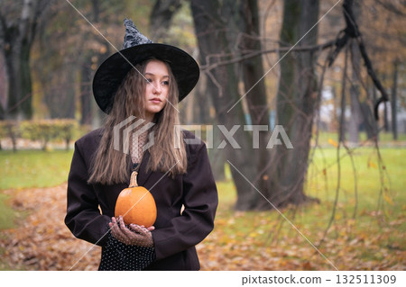 Teen girl in a witch hat holds an orange pumpkin close to the camera, creating a mystical autumn portrait with soft natural light Teen girl in a witch hat holds an orange pumpkin close to the camera, creating a mystical autumn portrait with soft natural light 132511309