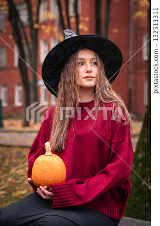 Teen girl in burgundy sweater and witch hat holds a pumpkin outdoors, surrounded by autumn leaves and brick buildings, creating a cozy Halloween vibe 132511311