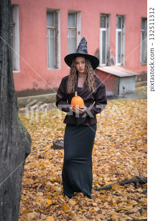 Teen girl in a witch hat and dark outfit holds an orange pumpkin outdoors, with autumn trees and pink brick buildings in the background 132511312