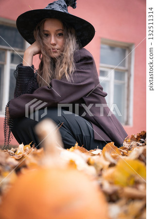 Teen girl in a witch costume sits among autumn leaves with a pumpkin in the foreground, posing against an old pink wall for a moody Halloween portrait 132511314