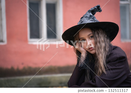 Teen girl in a witch hat poses thoughtfully against an old pink building, blending a moody Halloween atmosphere with urban charm Teen girl in a witch hat poses thoughtfully against an old pink building, blending a moody Halloween atmosphere with urban charm 132511317