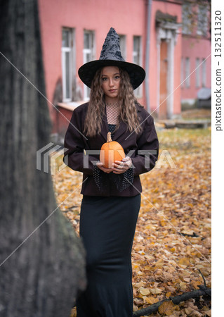 Teen girl in a witch hat and dark outfit holds an orange pumpkin outdoors, with autumn trees and pink brick buildings in the background 132511320