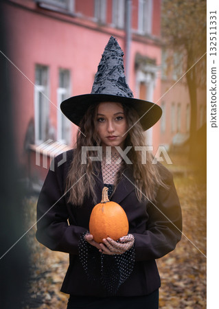 Teen girl in a witch hat and dark outfit holds an orange pumpkin outdoors, with autumn trees and pink brick buildings in the background 132511331
