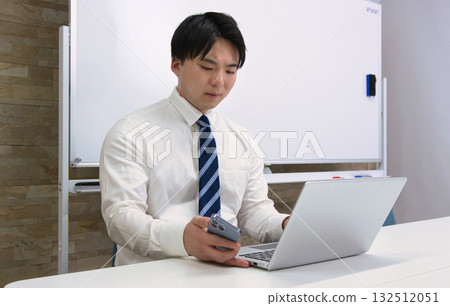 一位年輕的商人在辦公室裡一邊看著電腦一邊用智慧型手機聊天 一位年輕的商人在辦公室裡一邊看著電腦一邊用智慧型手機聊天 132512051