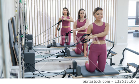 Three women in pink sportswear doing pilates exercises with a reformer bed.  132513100