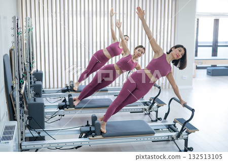 Three Asian women in pink sportswear doing side planks on a reformer. Pilates classes. Three Asian women in pink sportswear doing side planks on a reformer. Pilates classes. 132513105
