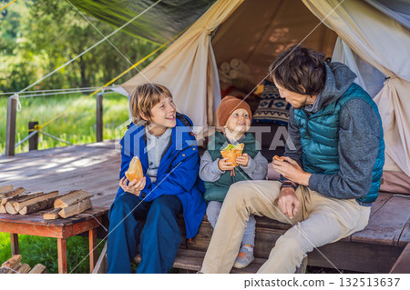 Father with his toddler and teenage son tourists in warm vests and hats eating buns while sitting on glamping steps, enjoying family travel, outdoor adventure, and bonding Father with his toddler and teenage son tourists in warm vests and hats eating buns while sitting on glamping steps, enjoying family travel, outdoor adventure, and bonding 132513637