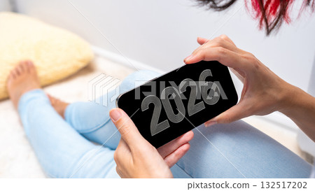 A young woman with red hair holds a smartphone displaying the year 2026. She sits on a light-colored floor, wearing blue jeans and relaxed attire. A young woman with red hair holds a smartphone displaying the year 2026. She sits on a light-colored floor, wearing blue jeans and relaxed attire. 132517202
