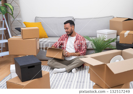A young man unpacks his belongings amidst carton boxes in his new home, a symbol of his relocation journey, whether renting or buying A young man unpacks his belongings amidst carton boxes in his new home, a symbol of his relocation journey, whether renting or buying 132517249