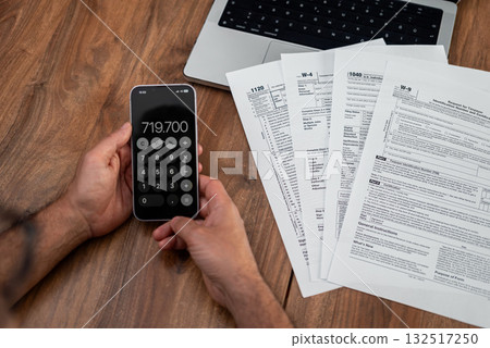 Man using a smartphone calculator with tax forms and a laptop on a wooden desk. Tax season, financial management, and accounting concept Man using a smartphone calculator with tax forms and a laptop on a wooden desk. Tax season, financial management, and accounting concept 132517250