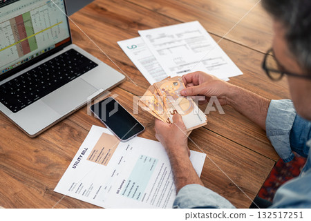 Man analyzing utility bills and counting euro cash at a wooden table with a laptop and smartphone. Household budget management and financial planning concept. Expense tracking and bill payment Man analyzing utility bills and counting euro cash at a wooden table with a laptop and smartphone. Household budget management and financial planning concept. Expense tracking and bill payment 132517251