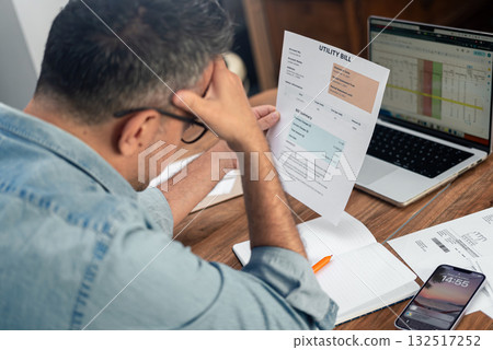Man holding utility bill with hand on forehead while working on budget at home. Concept of financial stress, household expenses, and bill management  132517252