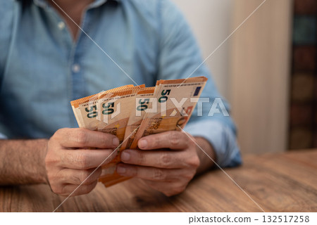Man holding multiple 50 euro banknotes in both hands on a wooden table. Financial concept highlighting cash handling, savings, and personal finance  132517258