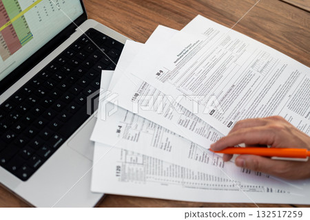 Close-up of accountant's hands reviewing tax forms with orange pen and laptop on wooden table. Financial planning and tax preparation concept Close-up of accountant's hands reviewing tax forms with orange pen and laptop on wooden table. Financial planning and tax preparation concept 132517259