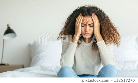 Young woman sitting on bed at home, suffering from strong headache caused by seasonal flu, feeling unwell, holding hands on temples, experiencing pain, fever and migraine, having health problems 132517270