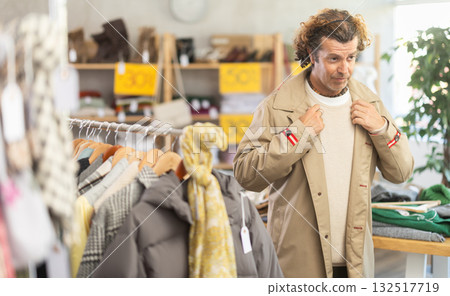 Interested man standing in clothing shop and trying new beige raincoat for cold season 132517719