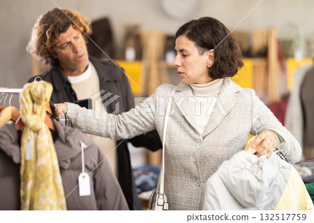 Woman choose jacket in store, exasperated husband accompanies his wife during shopping Woman choose jacket in store, exasperated husband accompanies his wife during shopping 132517759