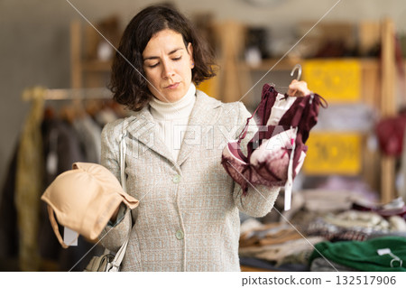 Focused female customer dressing in ordinary clothes holding bras in hands in underwear store 132517906