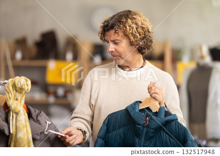 Stylish man standing in clothing shop and looking new down jacket made of high-quality material. Shopping and seasonal sales concept, winter season Stylish man standing in clothing shop and looking new down jacket made of high-quality material. Shopping and seasonal sales concept, winter season 132517948