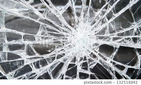 Close up of shattered glass revealing a complex web of cracks and fractures radiating from the impact point against a dark background, highlighting damage and vulnerability 132518842