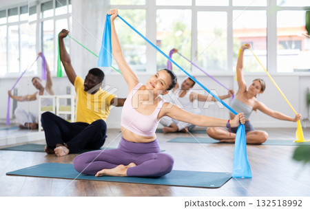 Group of multinational women and men sitting on sports mats and doing arm stretches using resistance bands at Pilates studio 132518992
