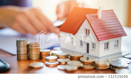 Miniature house surrounded by stacks of coins, representing mortgage payments, sits on a desk while an accountant calculates costs in the background Miniature house surrounded by stacks of coins, representing mortgage payments, sits on a desk while an accountant calculates costs in the background 132519200
