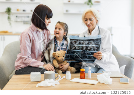 A doctor explains an X-ray to a mother and her young daughter, who is holding a teddy bear, in a cozy setting. A doctor explains an X-ray to a mother and her young daughter, who is holding a teddy bear, in a cozy setting. 132525935