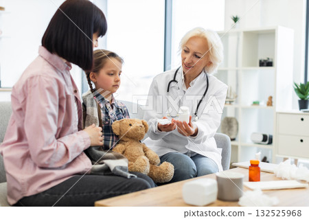A doctor is showing medicine to a young girl and her mother, likely explaining the dosage and use of the medication in a medical setting. 132525968