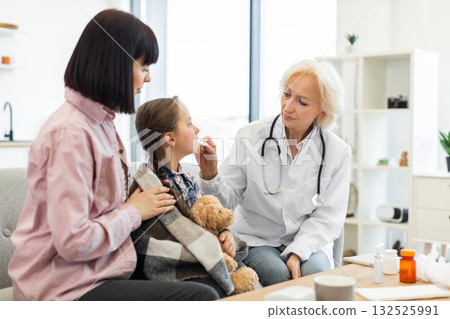 A female doctor checks a child's temperature while the mother comforts her in a doctor's office setting. The girl is wrapped in a blanket with a teddy bear. 132525991